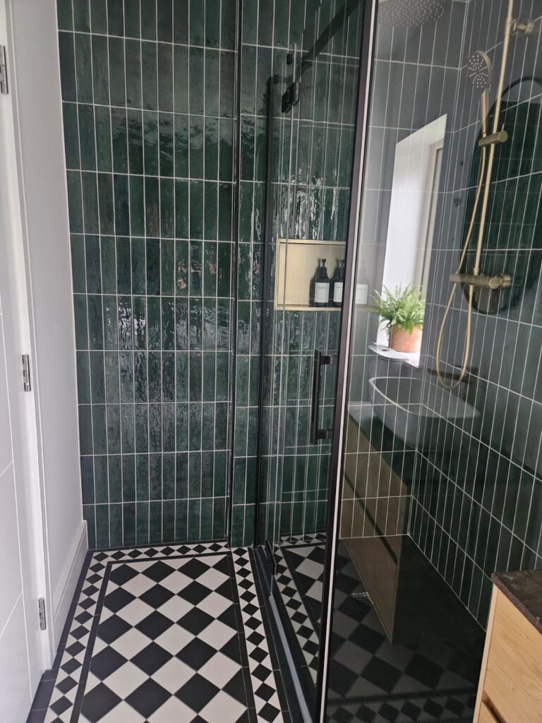Walk-in shower with green vertically stacked subway tiles, black-and-white Victorian patterned floor tiles, and brass shower fittings.