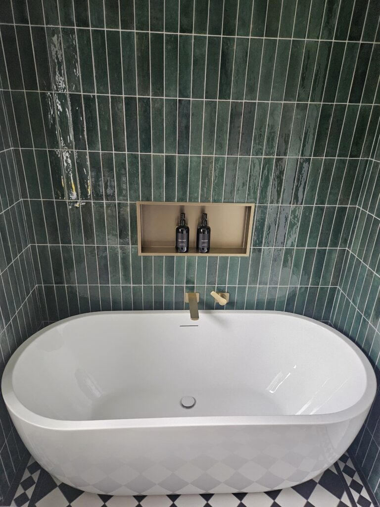 Freestanding white bathtub against vertically stacked green subway tiles with built-in niche and brass tap fittings.